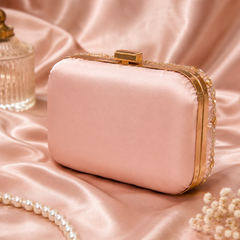A blush pink satin evening clutch bag with gold-tone hardware and a beaded trim, styled on a pink satin fabric background with decorative props.