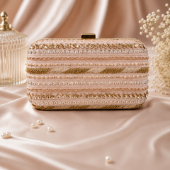 A rectangular evening clutch bag with ornate horizontal stripes of gold and silver beads, sequins, and faux pearls, resting on a draped satin fabric backdrop.