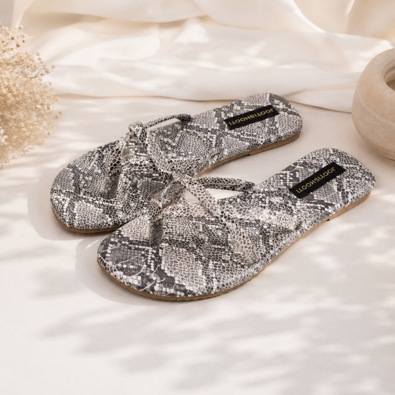 A pair of women's flat slide sandals with a gray and white snakeskin pattern, displayed on a soft white fabric background with decorative dried flowers and a ceramic bowl.