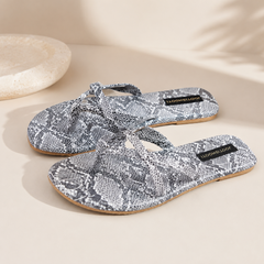 A pair of flat thong sandals with a gray and white snakeskin pattern, featuring a black brand label that reads 'LISANDRA LOUISE' on a warm beige background.