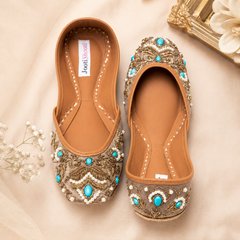 A pair of women's flat shoes with tan leather uppers and extensive gold embroidery, turquoise bead accents, and white faux pearls, displayed on a beige fabric background.