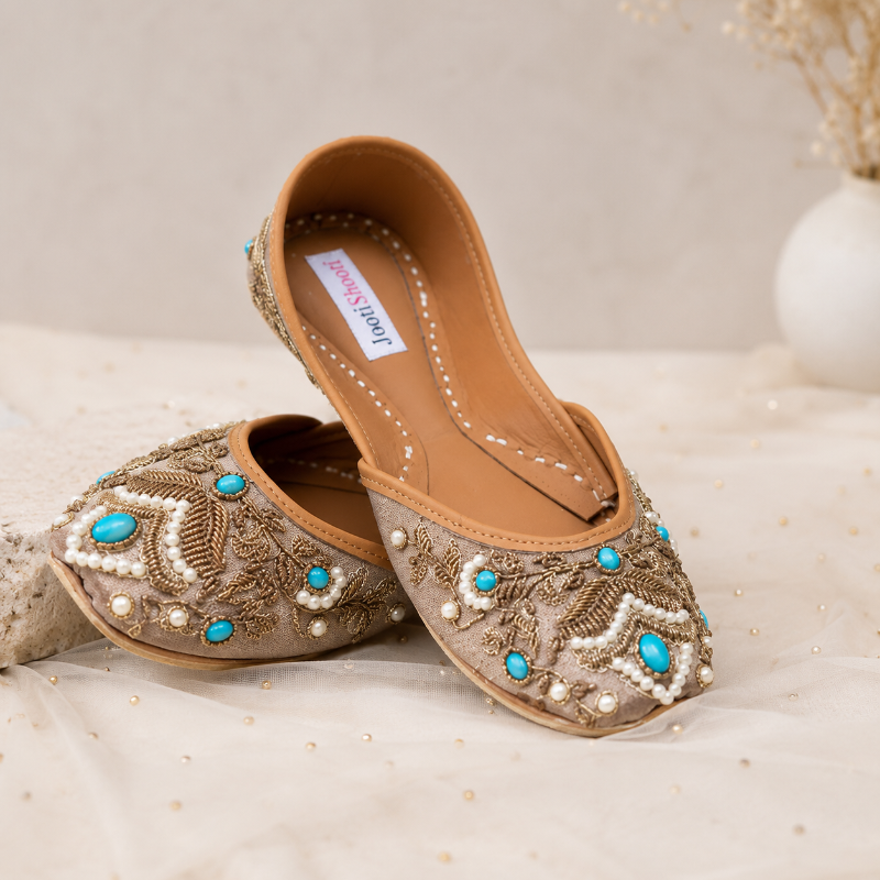 A pair of beige flat shoes with intricate gold embroidery, white pearl beadwork, and turquoise blue accents, displayed on a soft beige fabric surface.