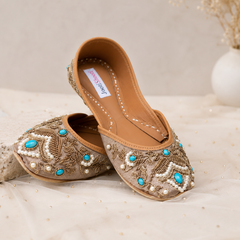 A pair of beige flat shoes with intricate gold embroidery, white pearl beadwork, and turquoise blue accents, displayed on a soft beige fabric surface.