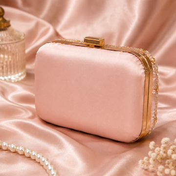 A blush pink satin evening clutch bag with gold-tone hardware and a beaded trim, styled on a pink satin fabric background with decorative props.