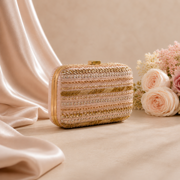 A rectangular evening clutch bag with ornate gold and beige horizontal beadwork and sequin detailing, styled on a beige surface with draped satin fabric and pink roses in the background.